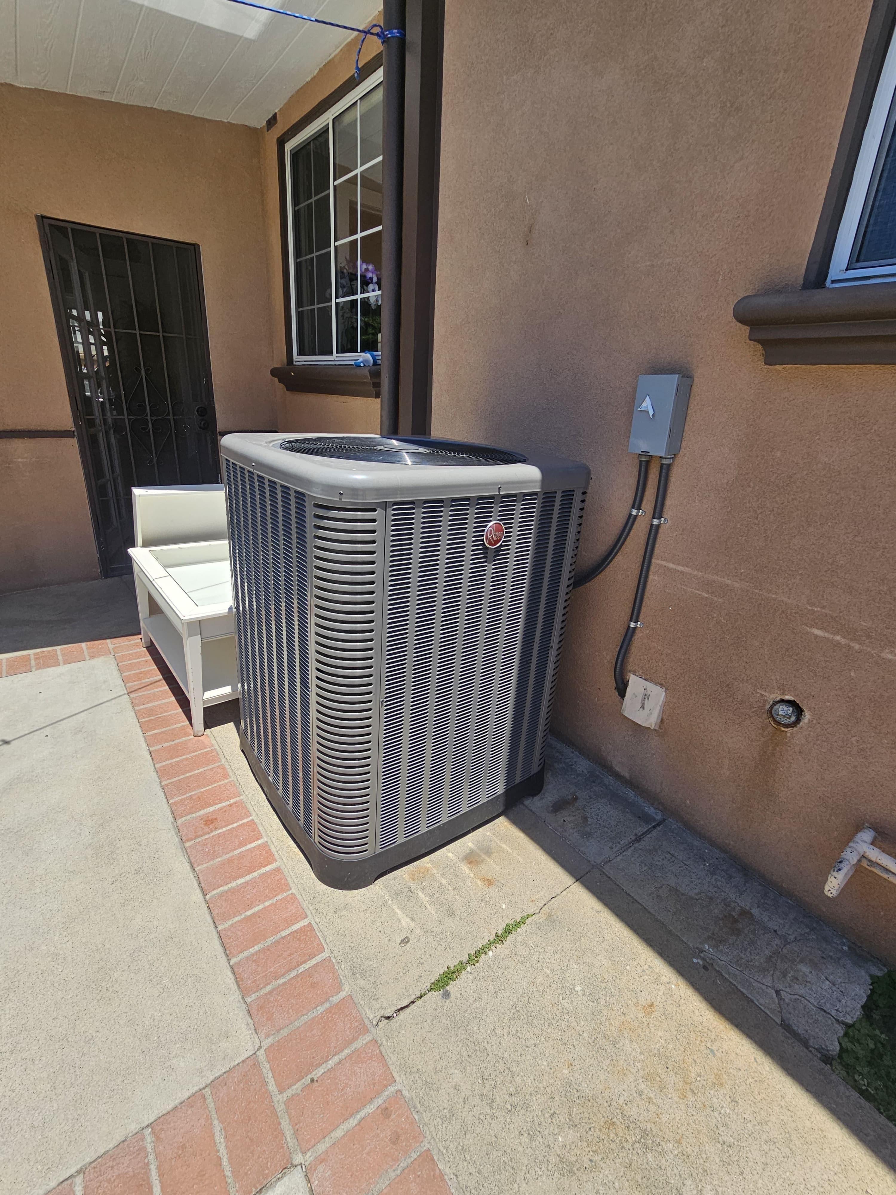 Condensador de AC Rheem instalado en un patio lateral angosto junto a la entrada de una vivienda